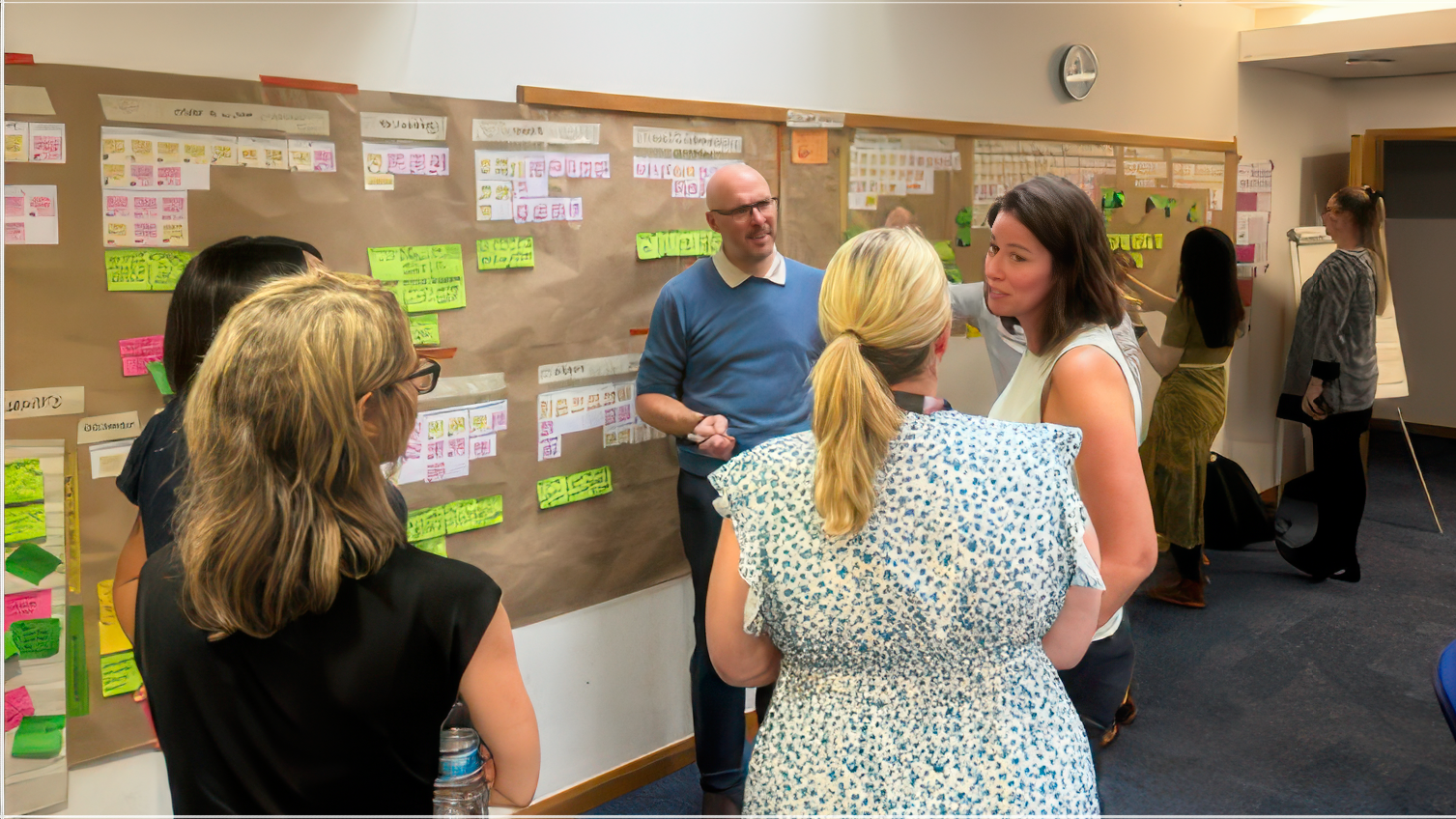 Group of people in a workshop setting, discussing ideas in front of large brown paper boards covered with sticky notes and organised cards on the wall